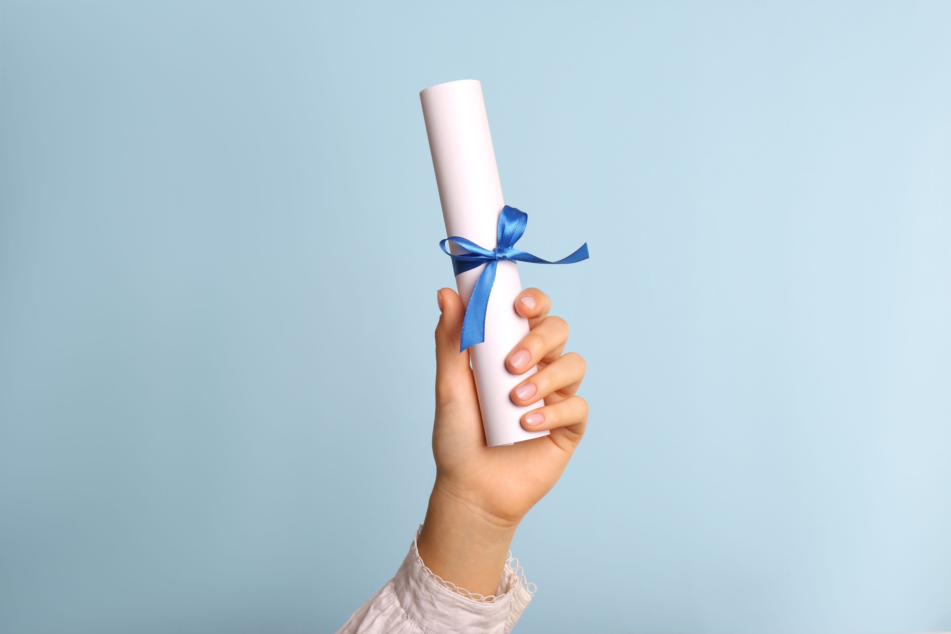 Student holding rolled diploma with ribbon on light blue background, closeup